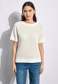 Pull en tricot blanc à manches courtes, présentant un motif en tricot ouvert et une texture lisse. Associé à un jean bleu.
