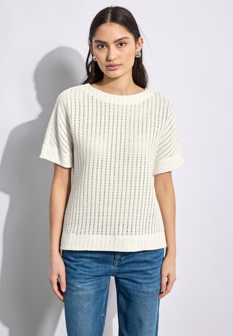 Pull en tricot blanc à manches courtes, présentant un motif en tricot ouvert et une texture lisse. Associé à un jean bleu.