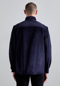 Camisa de terciopelo azul marino de manga larga con cuello alto y puños abotonados. Textura suave, corte relajado y detalle en el yugo trasero.