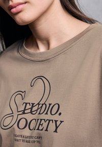 Khaki tričko s kulatým výstřihem, na kterém je černý text "STUDIO. SOCIETY," s podtitulem níže. Hladká tkanina s minimálními detaily šití.