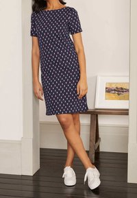 Marinblå klänning med korta ärmar, prydd med ett upprepat rosa mönster. Bärs med vita sneakers, mot en minimal bakgrund.