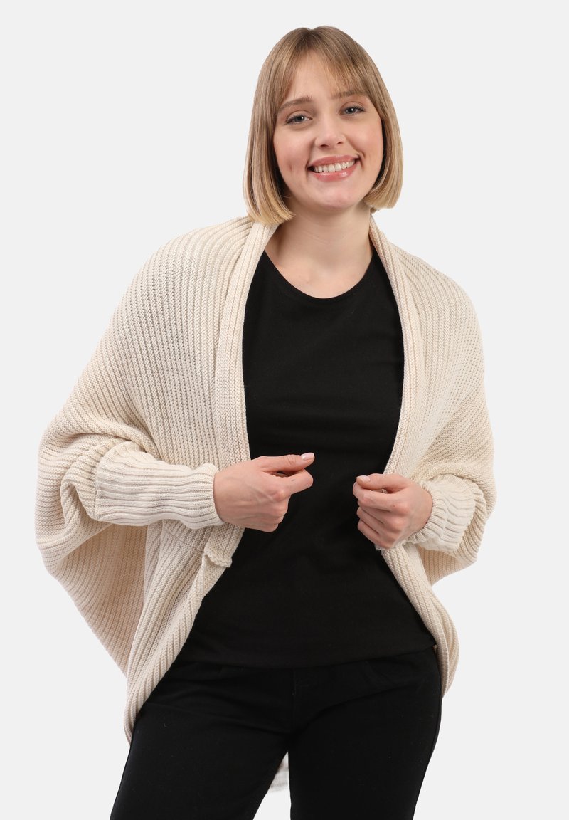 Femme souriant, portant un gilet en tricot beige sur une chemise noire et un pantalon noir, se tenant devant un fond blanc uni.