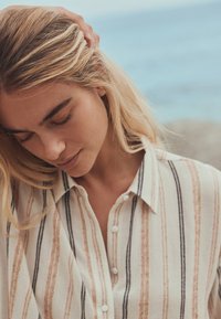 Young woman with closed eyes and blonde hair wears a striped button-up shirt, touching her hair with one hand, with a blurred blue background.