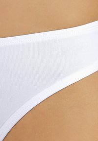 Witte bikinibroek met ribbeltextuur, voorzien van een gladde tailleband en nette stiknaden, getoond tegen een huiskleurige achtergrond.
