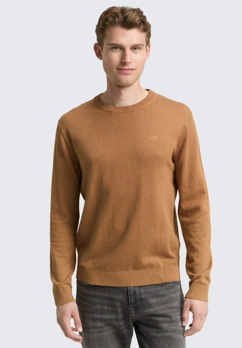 Pull en maille marron avec un col rond et des manches longues. Présente un logo discret sur la poitrine et des bords côtelés au niveau de l'ourlet et des poignets.