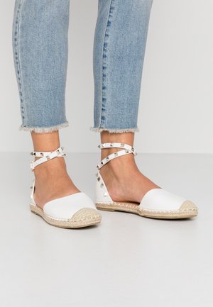 Hvide espadriller med flade såler, ankelstropper og sølvnitter, båret med lyseblå jeans med flossede kanter på en neutral baggrund.
