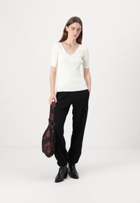 Rippendes weißes kurzärmeliges Oberteil mit V-Ausschnitt, kombiniert mit schwarzen lockeren Hosen. Das Model hält eine braune Batik-Schultertasche. Schwarze Knöchelstiefel.