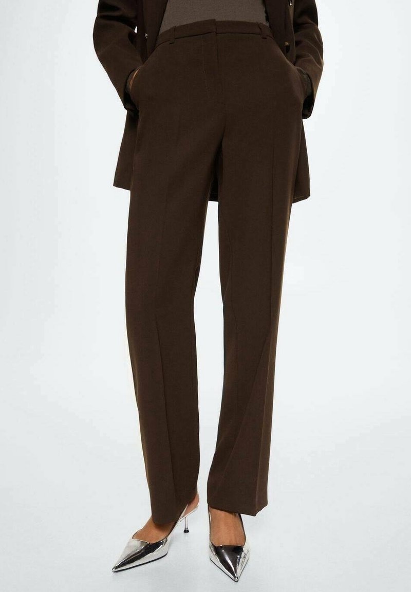 Personne portant un pantalon marron foncé sur mesure et des chaussures à talons hauts argentées pointues, debout, les mains dans les poches, sur fond blanc.