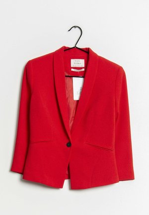 Czerwona, dopasowana marynarka z jednym czarnym guzikiem, dwoma przednimi kieszeniami z rozcięciami, wisząca na czarnym wieszaku na białym tle.