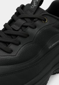 Svart idrottssko med strukturerad yta, snörning och tjock gummisula. Har varumärkesdetaljer och släta detaljer.