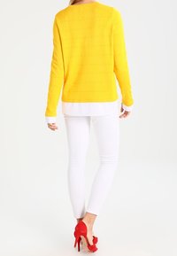 Maglione giallo a maglia con righe orizzontali, strato sottostante bianco, jeans skinny bianchi e scarpe con tacco rosso.