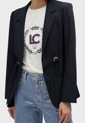 Vrouw draagt een zwart blazer met trekkoorddetails, een wit bedrukt T-shirt en blauwe spijkerbroek met pailletten.