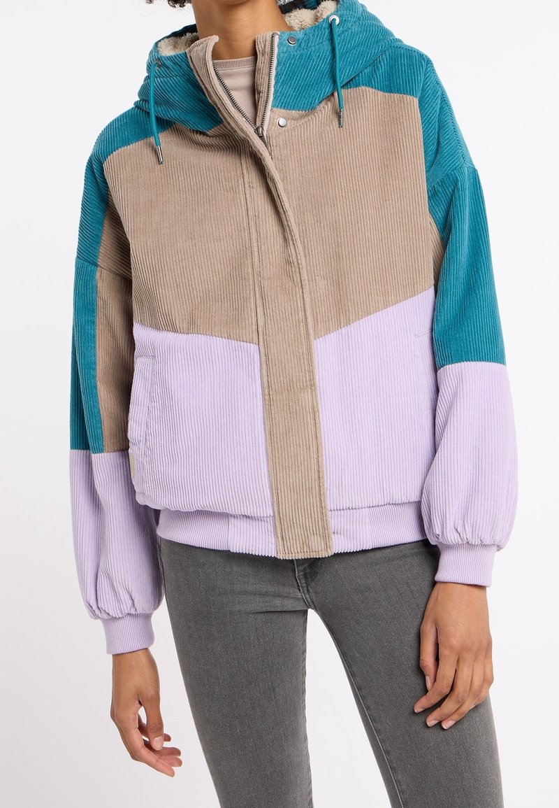Personne porte une veste à capuche en velours côtelé avec des blocs de couleur turquoise, beige et violet clair et un pantalon gris ajusté sur fond blanc.