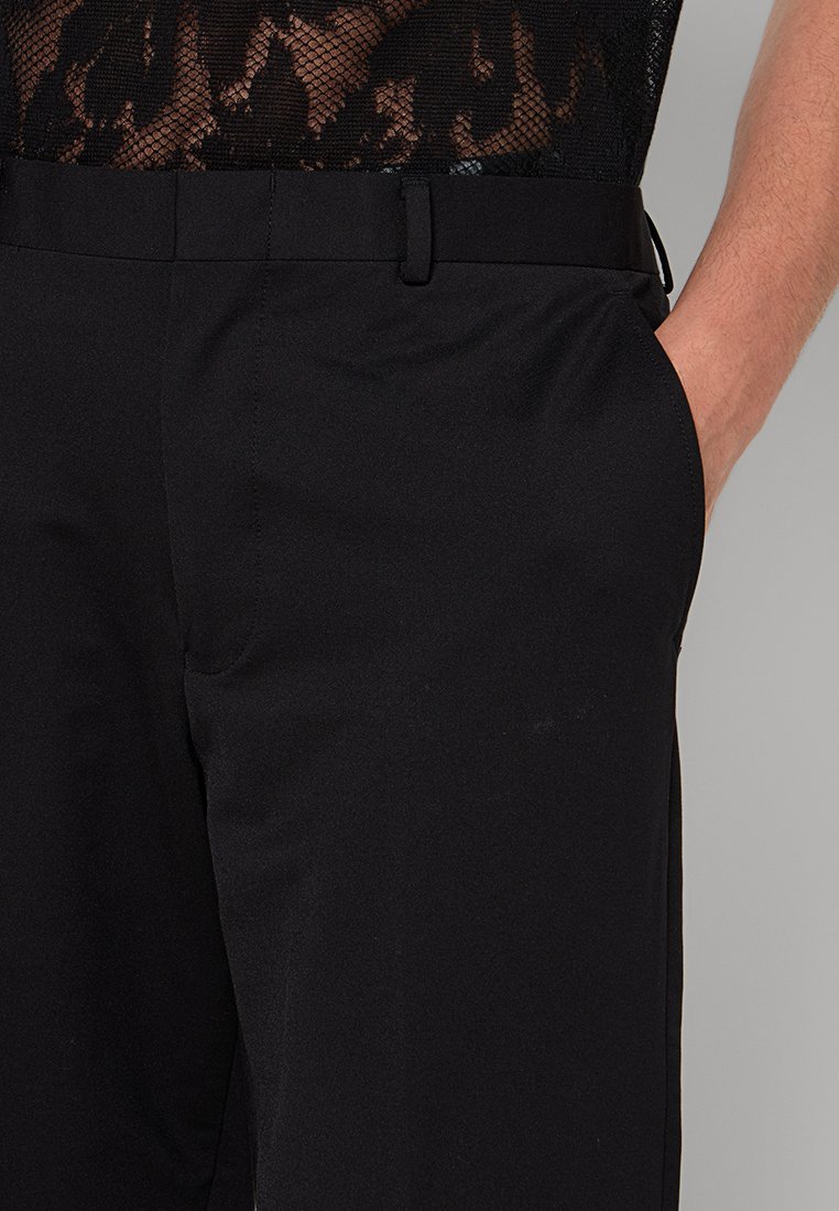 Primo piano di pantaloni neri indossati da una persona, caratterizzati da una cintura sottile e una maglia in pizzo trasparente sottostante, che mette in evidenza un mix di trame.