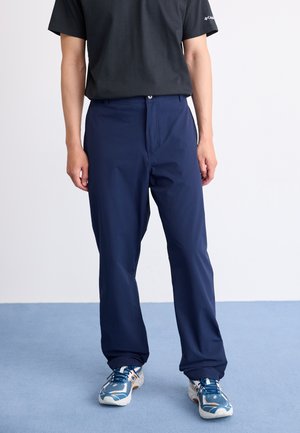 Man in een marineblauwe broek, zwart t-shirt en blauw-witte sneakers staat op een lichtblauwe tapijt tegen een witte muur.