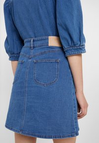 Denimová sukně v středně modré barvě. Má dvě zadní kapsy, viditelný pas s koženou etiketou a mírně rozšířený lem.