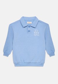 Ανοιχτό μπλε πουλόβερ με γιακά πόλο, τρία κουμπιά και μακριά μανίκια. Διαθέτει λευκό λογότυπο με το γράμμα «Α» στο αριστερό στήθος. Μαλακή υφή.