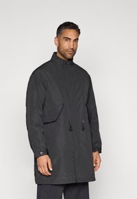 Black long jacket with high collar, front snap closure, adjustable drawstrings, and side pockets. Made from a lightweight, textured fabric.