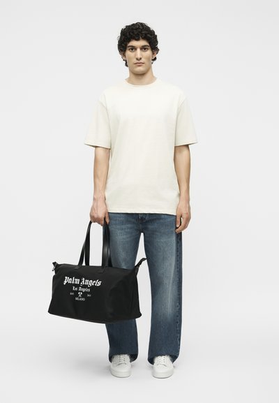 Νέος άνδρας με λευκό μπλουζάκι και μπλε τζιν κρατάει μια μαύρη σάκα Palm Angels με λευκό λογότυπο, στέκεται μπροστά σε απλό λευκό φόντο.