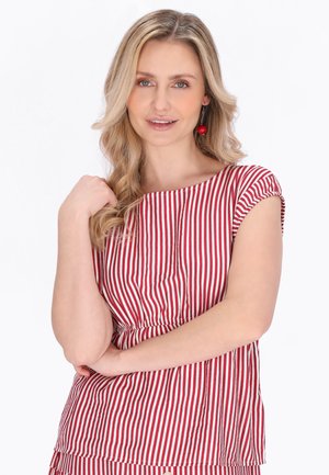 Lyshåret kvinde med bølget hår iført en top med røde og hvide lodrette striber og røde kirsebærformede øreringe, der smiler blidt med armene krydsede.