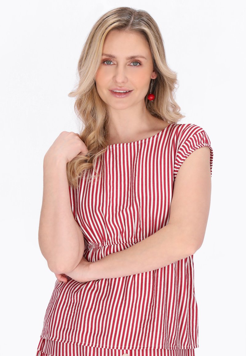 Blondýnka s vlnitými vlasy má na sobě červenobíle pruhované tričko a červené náušnice ve tvaru třešní, jemně se usmívá a má založené ruce.