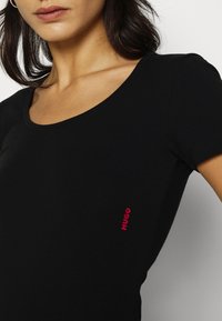 T-shirt en coton noir avec un décolleté arrondi et des manches courtes. Présente un petit logo rouge "HUGO" sur la poitrine gauche. Texture lisse.
