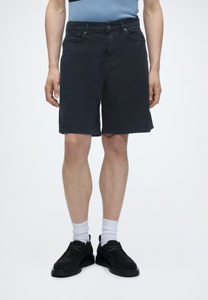 Homme portant un short noir au genou, des chaussettes blanches et des chaussures noires à lacets, debout devant un fond blanc uni.