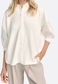 Frau in einer off-white, lockeren Bluse mit dreiviertel Puffärmeln und beigen, weiten Hosen, mit der Hand in der Tasche, stehend vor einem einfarbigen Hintergrund.