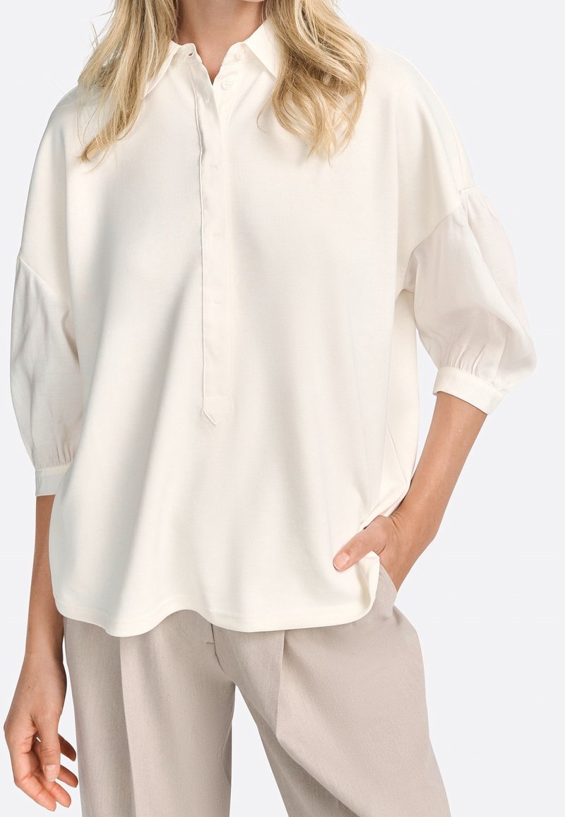 Frau in einer off-white, lockeren Bluse mit dreiviertel Puffärmeln und beigen, weiten Hosen, mit der Hand in der Tasche, stehend vor einem einfarbigen Hintergrund.