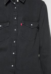 Camicia in denim nero con chiusura a bottoni, due tasche sul petto, bottoni argentati e un'etichetta con il logo rosso sulla tasca sinistra. Tessuto testurizzato.