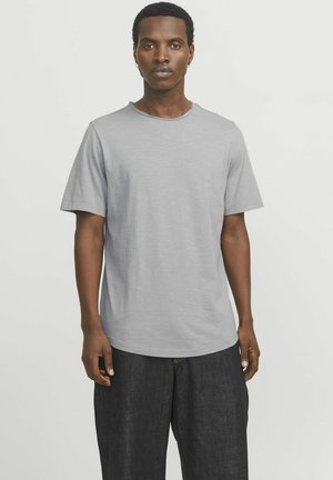 Camiseta gris claro de manga corta, con escote redondo y un acabado ligeramente texturizado, combinada con pantalones de mezclilla oscuros.
