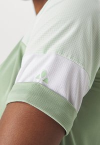 Vaaleanvihre ä urheilupaita, jossa on teksturoitu kuvio, kontrastivärinen valkoinen hiha ja pieni logokoriste hihassa.