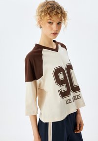 Camiseta de manga corta y escote en V en color crema y marrón, con un gran gráfico de "90" y el texto "LOS ALES", hecha de suave tela de algodón.