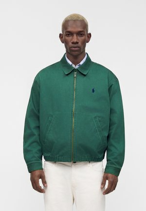 Homme aux cheveux blonds courts portant une veste verte zippée par-dessus une chemise bleu clair à col et un pantalon blanc sur un fond uni.