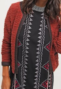Kvinna som bär en texturerad röd kofta över en svart klänning prydd med geometriska mönster i vitt och rött. Håret faller i mjuka vågor.