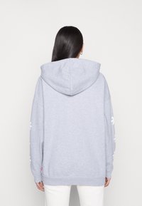 Sudadera gris con una gran capucha, corte oversized y diseños florales blancos en las mangas. Presenta un dobladillo acanalado y una textura de tela suave.