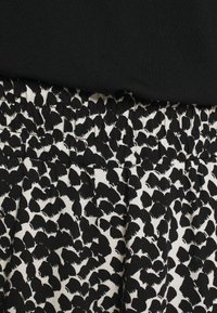 Close-up van een zwarte elastische tailleband met geplooide stof in een zwart-wit abstract bladmotief onder een effen zwarte top.