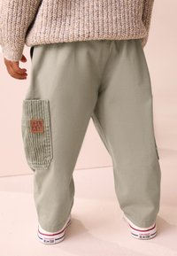 Pantaloni cargo verde chiaro con tasca a coste e etichetta marrone con scritta "PLAY". Abbinati a scarpe da ginnastica bianche con dettagli rossi e blu.