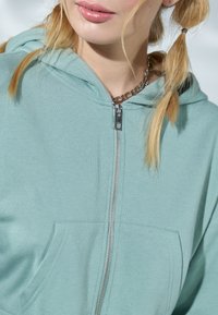 Sudadera de cremallera verde claro con un bolsillo frontal, textura suave y capucha ajustable. Detalle en cremallera plateada y un diseño simple y limpio.
