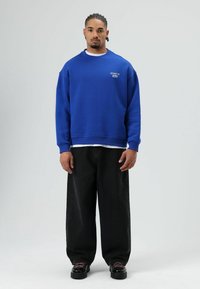 Sweat-shirt bleu à col rond avec poignets côtelés, superposé sur un t-shirt blanc, associé à un jean noir à jambes larges et des chaussures noires épaisses.