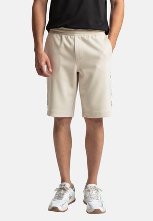 Hombre con pantalones cortos beige hasta la rodilla, camiseta negra y zapatillas blancas, de pie con las manos dentro y junto a los bolsillos sobre fondo blanco.