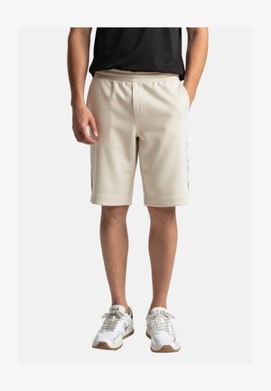 Uomo con pantaloncini beige fino al ginocchio, top nero e scarpe da ginnastica bianche, in piedi con le mani nelle tasche e accanto alle tasche su sfondo bianco.