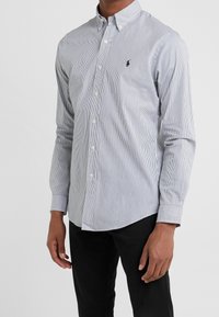 Mężczyzna ubrany w biało-niebieską koszulę w prążki z długim rękawem oraz czarne spodnie, stojący na jednolitym tle.