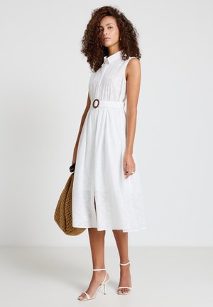 Donna con capelli ricci che indossa un vestito midi bianco senza maniche con cintura e tacchi bianchi, che tiene una grande borsa intrecciata marrone.