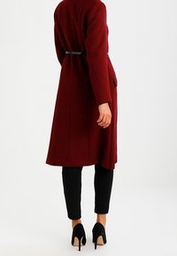 Donna che indossa un lungo cappotto bordeaux, pantaloni neri aderenti e scarpe con tacco nere, in piedi contro uno sfondo bianco semplice.