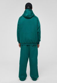 Teal fleece hoodie met capuchon en buidelzak, gecombineerd met bijpassende wijdvallende broeken. Materialen lijken zacht met zichtbare stikdetails.