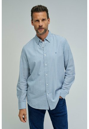 Hombre con camisa de botones a rayas verticales en azul claro y blanco, y pantalones azul oscuro, posando con una mano en el bolsillo.