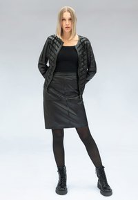 Vrouw met steil blond haar, gekleed in een zwarte leren jas, zwart topje, zwarte leren rok tot op de knie, zwarte panty's en zwarte veterlaarzen.