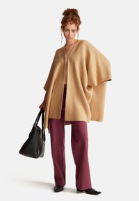 Velký cardigan v barvě velbloudí vyrobený z jemného materiálu, s hlubokým V výstřihem a otevřeným předním dílem. V kombinaci s burgundskými širokými kalhotami a černou kabelkou.