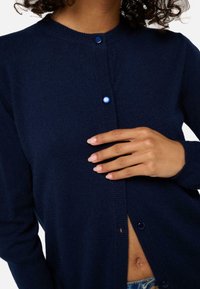 Cardigan bleu marine avec un col rond, comportant deux boutons bleus, des poignets côtelés et une texture douce, partiellement ouvert pour révéler la peau en dessous.
